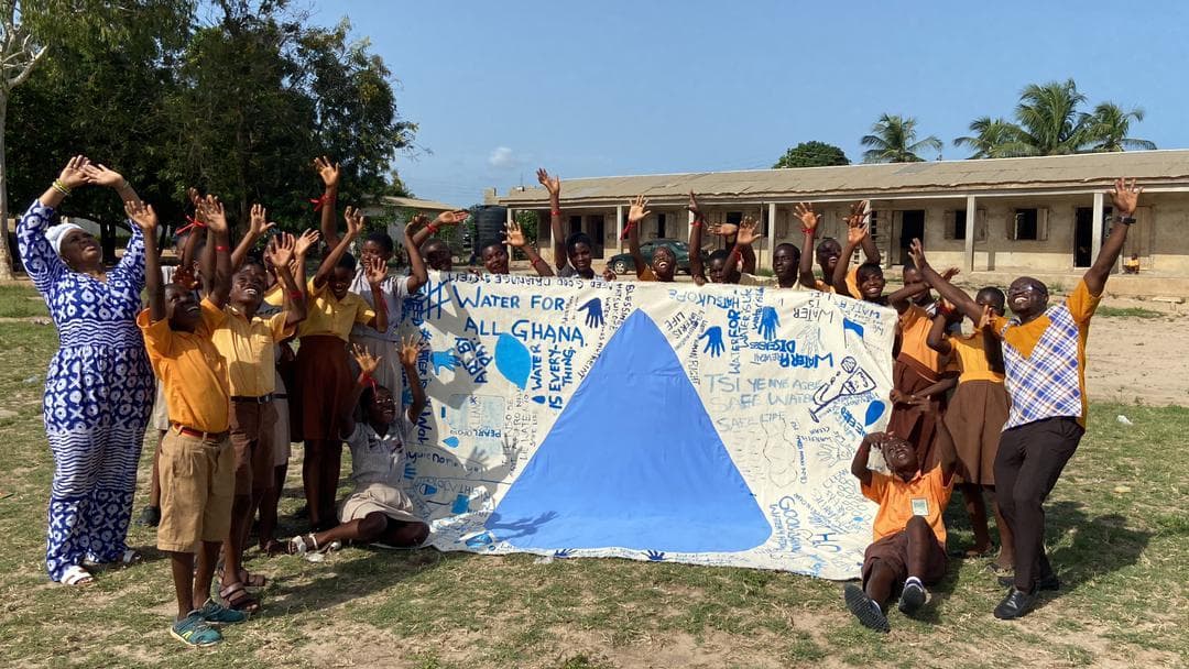 Ghanaian schoolchildren cheering behind a hand-painted "Water for All Ghana" banner